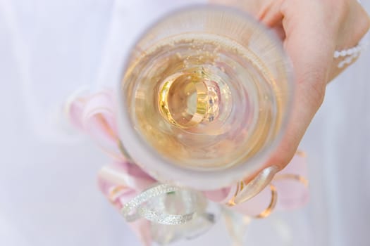 a hand of a bride holds a glass full of champagne and twwo gold rings in it