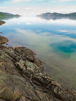 Calm summer day at rocky shore of Lake Laberge, Yukon Territory, Canada, reflecting sky and distant shoreline on mirror-like surface.