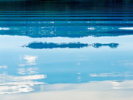 Distant shoreline, clouds and blue sky mirrored on calm lake surface with distortions of some ripples.