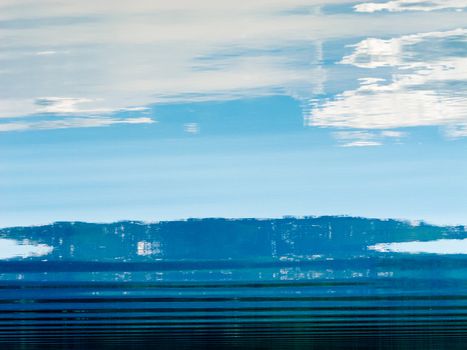 Distant shoreline, clouds and blue sky mirrored on calm lake surface with distortions of some ripples.