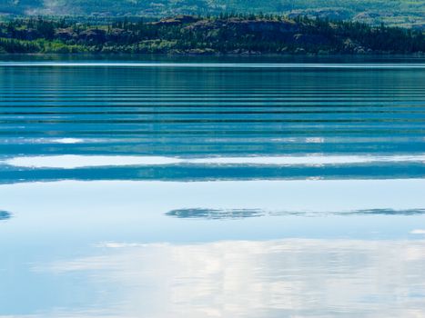 Distant shoreline, clouds and blue sky mirrored on calm lake surface with distortions of some ripples.