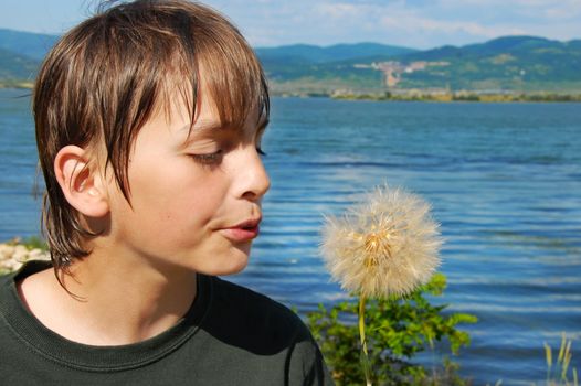 boy on riverbank blowing on dandelion flower portrait over rural landscape