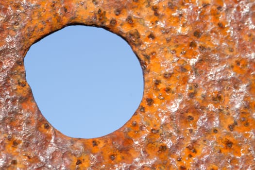 Background of rust, coloured red, orange and yellow with an irregular circle, hole with blue sky beyond.