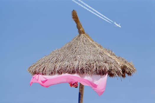 A sun shade of straw against a clear blue sky with an airplane painting white trails, suggesting the consept of summer tourism.