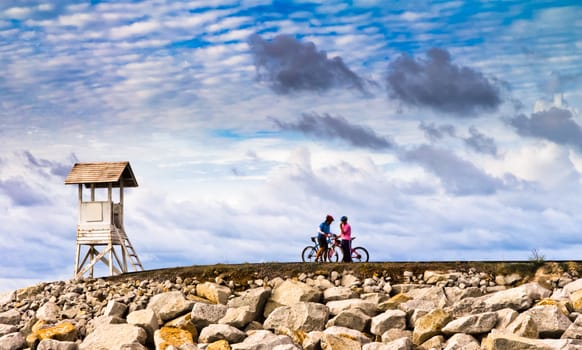 Men are towed bicycle and lighthouse at sunset.