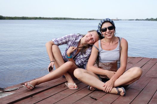 Beauty smiling teen girl and her mother sitting on wooden moorage near river