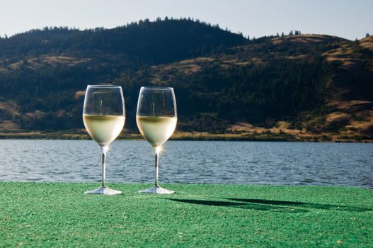 tow glasses of white wine sitting on the end of a dock by the lake