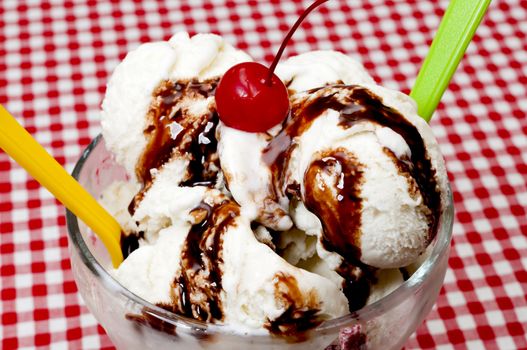 Vanilla ice cream and spoons with cherry and chocolate fudge topping on table with red gingham table cloth.