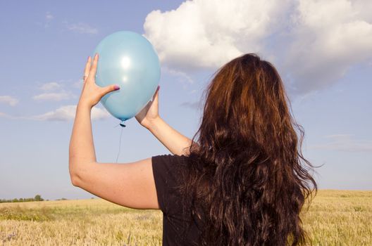 Young woman, dressed with black dress, holding blue balloon in the wheat field.