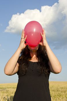 Young woman, dressed with black dress, holding red balloon in front of her face.