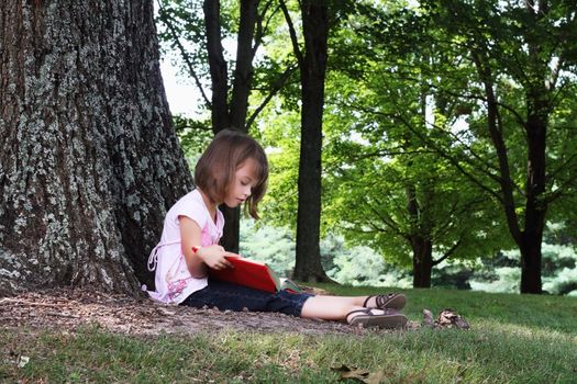 Little girl sits outdoors under a large oak tree and reads a book.
