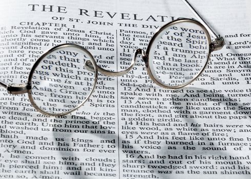 Old fashioned round reading glasses laying on a page from the bible on the revelation with strong shadow