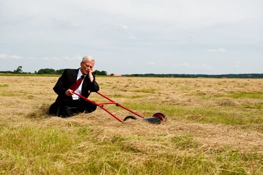 An exhausted businessman rest on his knees with his lawnmower in a field of harvested grain as he prepares to reap the rewards of all his hard labour and perseverance, conceptual image