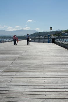 A group of people walks down the board walk of a pier with mountains in the distance.