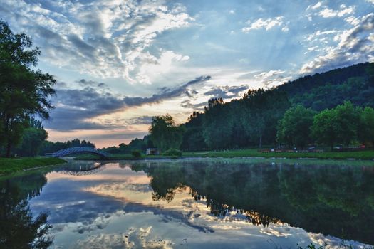 Beautiful sunrise and cloudscape at the side of a small fishing lake situated in a leisure park. The calm water surface magnificiently reflects the amazing colors of the early morning sky.