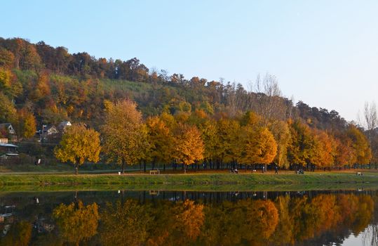 Small fishing lake in autumn, lit by the mellow late afternoon sunlight. Colors of foliage and reflections on the calm water surface are beautiful, yet create a gloomy atmosphere. Residents of the nearby town take a rest on the lakeside fishing, or just relaxing.