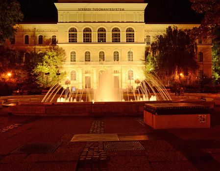 Beautifully lit fountain at night on Dugonics Square in downtown Szeged, Hungary. In the background, the building of the University of Szeged can be seen.