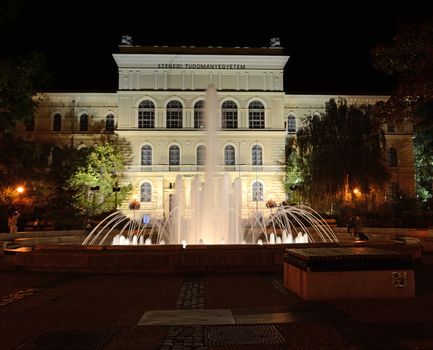 Beautifully lit, glowing fountain at night on Dugonics square in downtown Szeged, Hungary. In the background, the building of the University of Szeged can be seen.