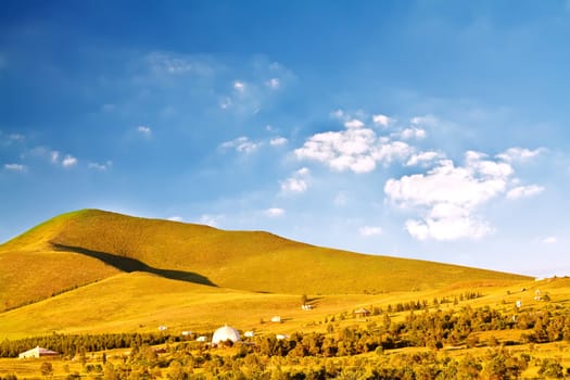 Landscape photo of plain with colorful grass under blue sky, with small huts aside a road along the hill