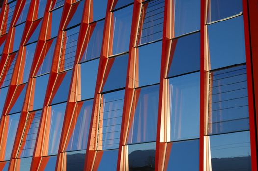 Detail of the front of an orange office block with windows reflecting the clear blue sky.