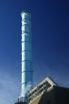 An industrial chimney, with cooling fins, and pipe-work, against a clear blue sky. Only a small amount of smoke being produced, from a small chimney below it.