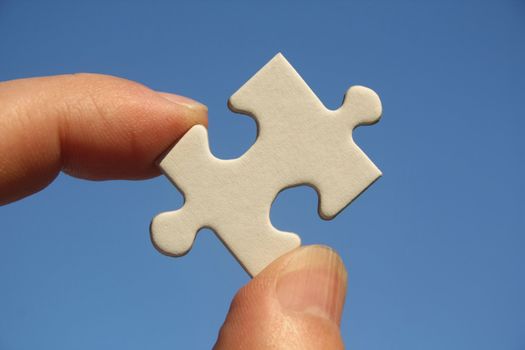 One white jigsaw puzzle piece in human fingers on a blue sky background
