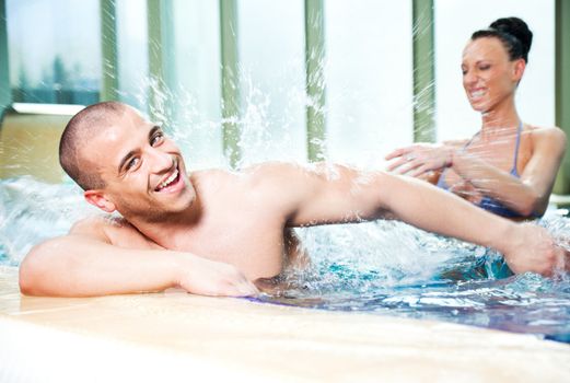 Young attractive couple having fun splashing in a swimming pool, man on focus