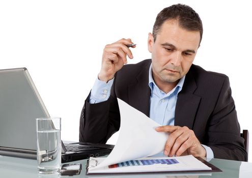 businessman working on desk reading documents by laptop, holding pen in hand