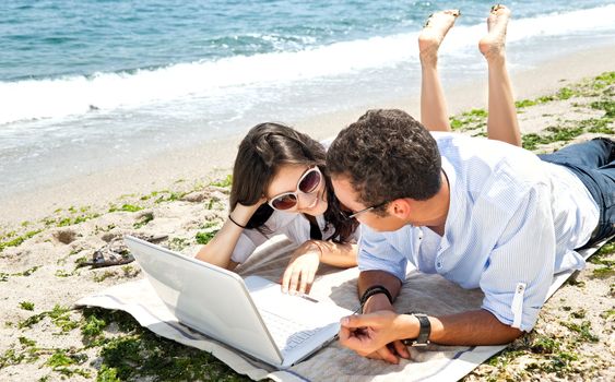 Young couple lying on the beach behind laptop