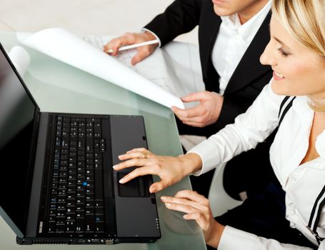 Two bussines persons, engineers, architects sitting on desk with blueprints and laptop