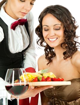 Young waitress offering fresh sliced fruits on wooden cutting board to beautiful female