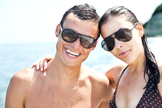 Happy couple with sunglasses hugging and smiling on beach, female with tongue out