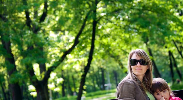 Mother with sunglasses sitting on bench in park embracing daughter