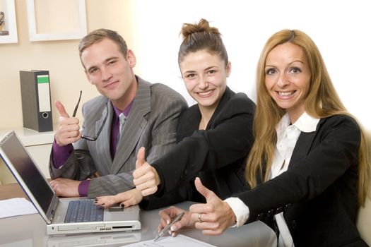 Positive team in the office shows up the thumb. Two women and a man sitting in front of a computer and smiling. Horizontal format and text space