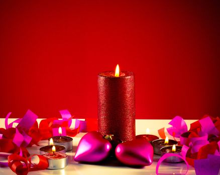 Burning candles with two heart shaped toys against red background