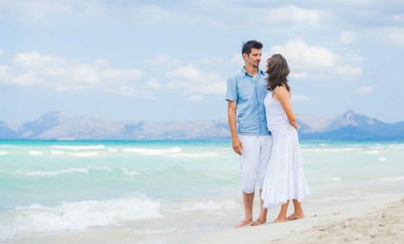 Happy young man and woman couple walking, laughing and holding hands on a deserted tropical beach with bright clear blue sky