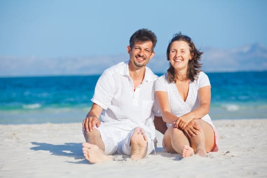 Happy young man and woman couple laughing and sitting on a deserted tropical beach with bright clear blue sky
