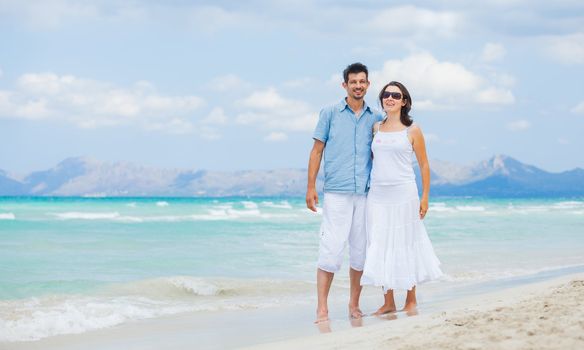Happy young man and woman couple walking, laughing and holding hands on a deserted tropical beach with bright clear blue sky