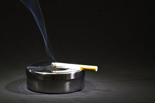 a cigarette burns on a metal ashtray with black background