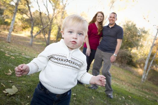 Cute Young Boy Walking in the park as Adoring Parents Look On From Behind.