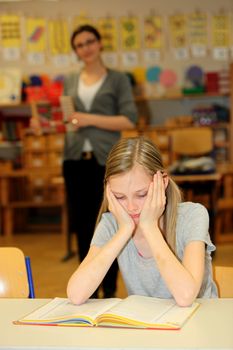 stressed-out student in the school with a book in front of. In the background is the teacher.