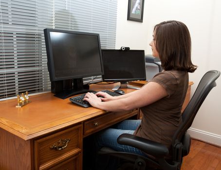 Young woman in home office with two monitors and keyboard on leather desk and typing on keyboard