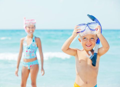 Happy young boy with snorkeling equipment on sandy tropical beach, his sister background.