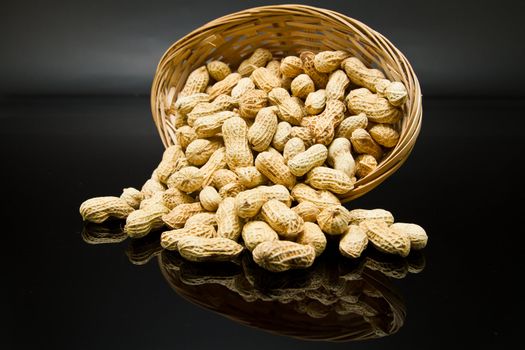 A pile of roasted groundnut spilled from a rattan basket on a black mirror with mirror image