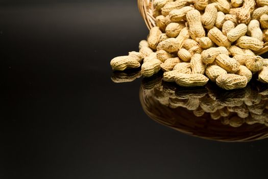 A pile of roasted groundnut spilled from a rattan basket on a black mirror with mirror image off-centered to the top right