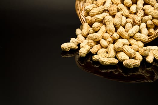 A pile of roasted groundnut spilled from a rattan basket on a black mirror with mirror image off-centered to the top right leaving empty space for text