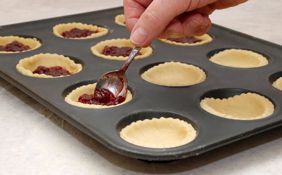 A woman's hand holding a teaspoon as she fills jam tarts with raspberry conserve