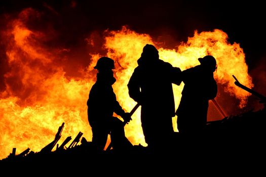 Three firemen fighting a raging fire with huge flames of burning scrap timber