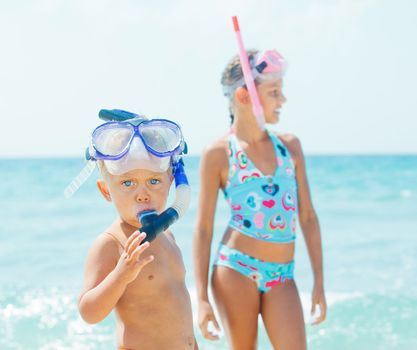 Happy young boy with snorkeling equipment on sandy tropical beach, his sister background.