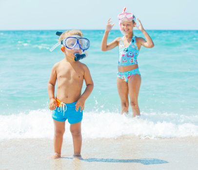 Happy young boy with snorkeling equipment on sandy tropical beach, his sister background.